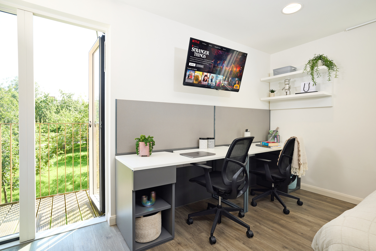 Modern home office with two black chairs at a shared white desk. A TV on the wall displays a streaming service interface. Shelves with plants and decor are above the desk. A door opens to a balcony with a view of greenery.