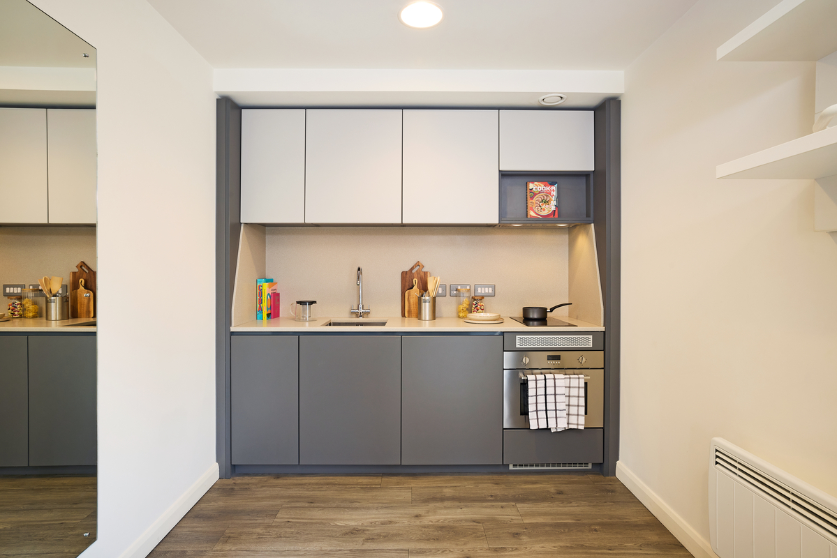 Modern kitchen with gray and white cabinets, a stainless steel sink, and a stove with a towel hanging. Wooden flooring, various kitchen items on the counter, and a shelf with pantry items add warmth and functionality.