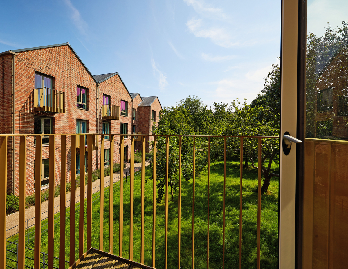 View from a balcony showing modern red-brick apartment buildings with small balconies, surrounded by a lush green garden. The sky is clear and blue, and a pathway is visible through the greenery.