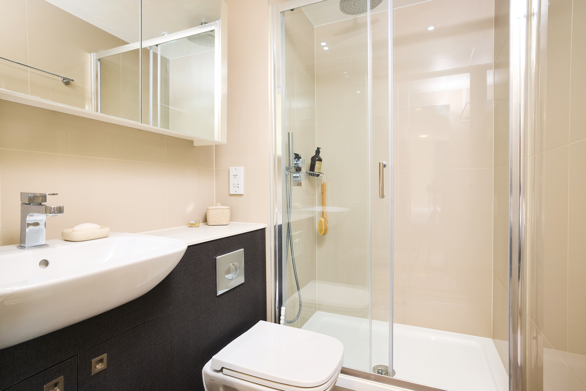 A modern bathroom with a glass-enclosed shower, white sink with a rectangular basin, and a toilet. The shower has a rainfall showerhead and a hanging sponge. Neutral-toned walls and a mirrored cabinet above the sink complete the look.