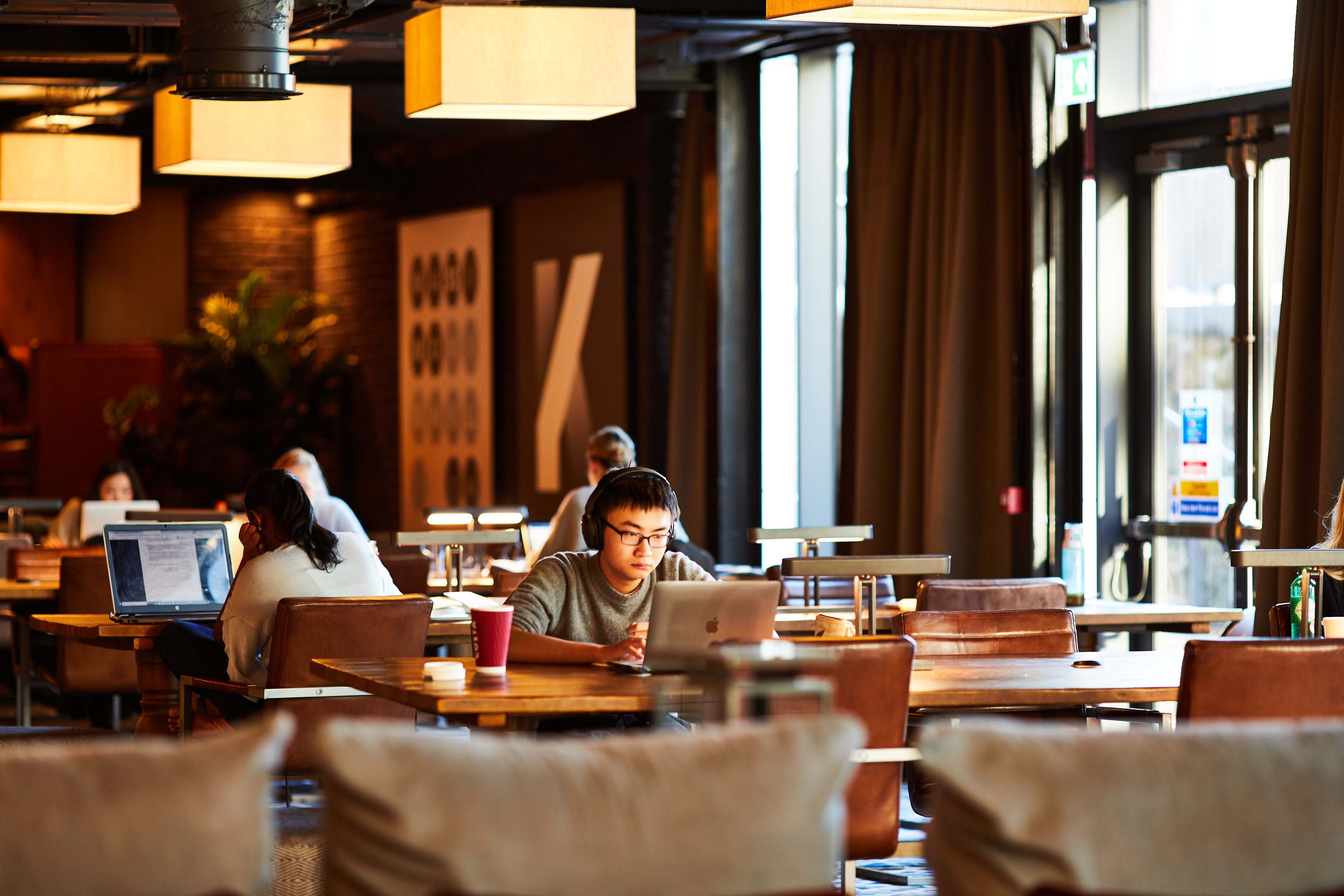 A spacious coffee shop with warm lighting serves as an ideal study space, where several people sit at wooden tables working on laptops. In the foreground, a person wearing glasses focuses intently on their screen, with a coffee cup nearby—a perfect setting for gaining academic advantage.