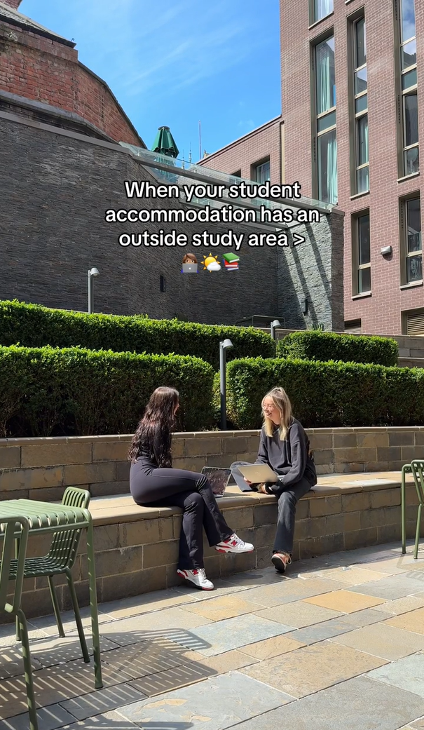 Two people are sitting on a stone bench in an outdoor study area, surrounded by neatly trimmed hedges and buildings. One is using a laptop. The sky is clear and sunny. Text on the image reads, "When your student accommodation has an outside study area.