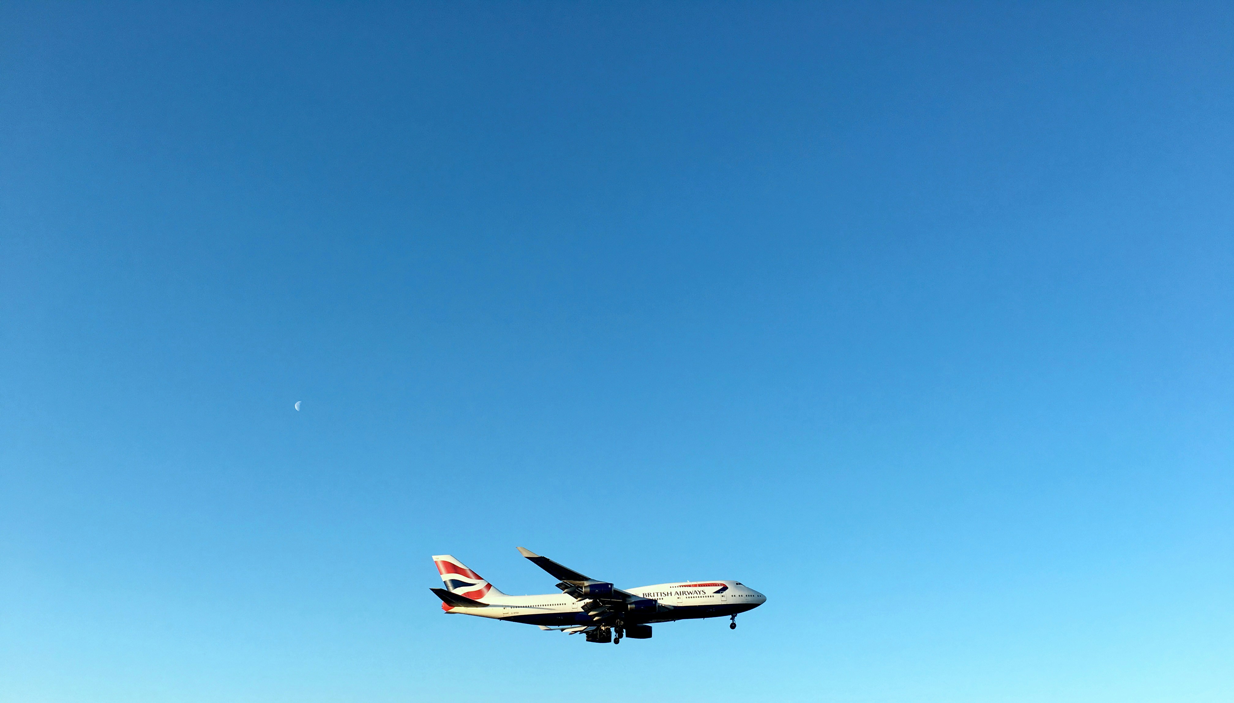 An airplane soars against a clear blue sky, gracefully arcing past a small crescent moon, as if charting its course to Exeter Airport.