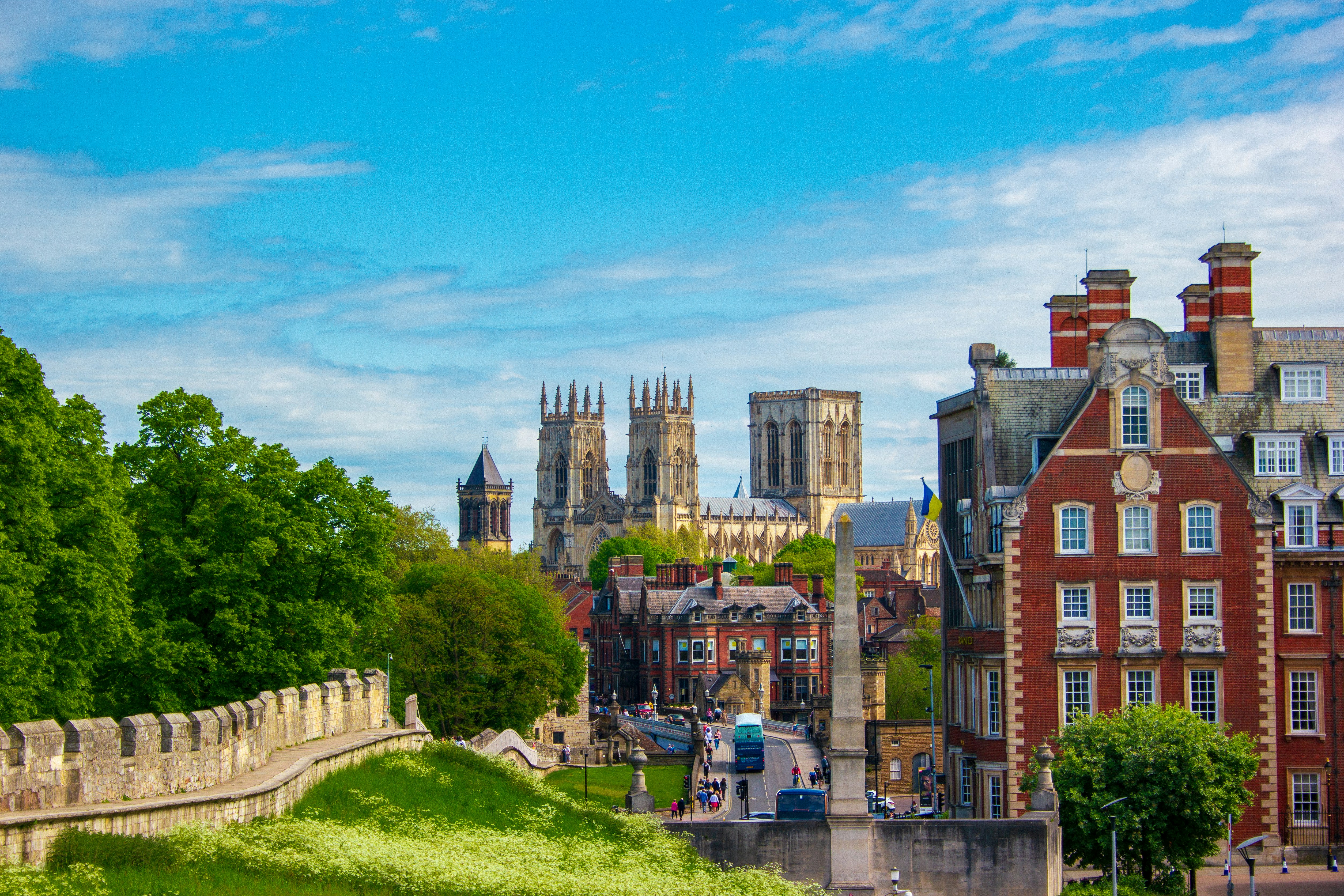 A scenic view of the historic city of York features a grand cathedral with multiple towers in the background. The foreground includes a stone wall and red brick buildings, surrounded by lush greenery under a partly cloudy blue sky—a perfect setting to study or simply enjoy life's moments.