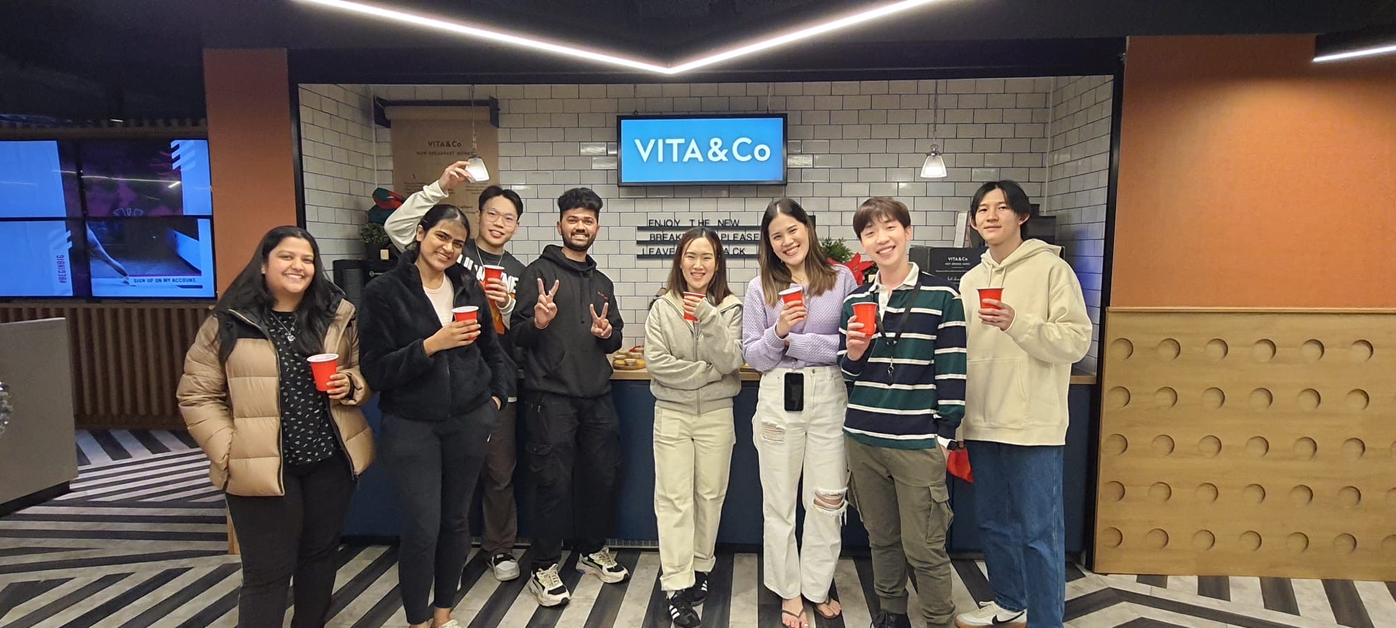 A group of nine people, part of the Vita Student network, stand in a room with a "VITA & Co" sign on the wall. They hold red cups and smile, with some making peace signs. The striped floor and tiled wall add to the lively ambiance.