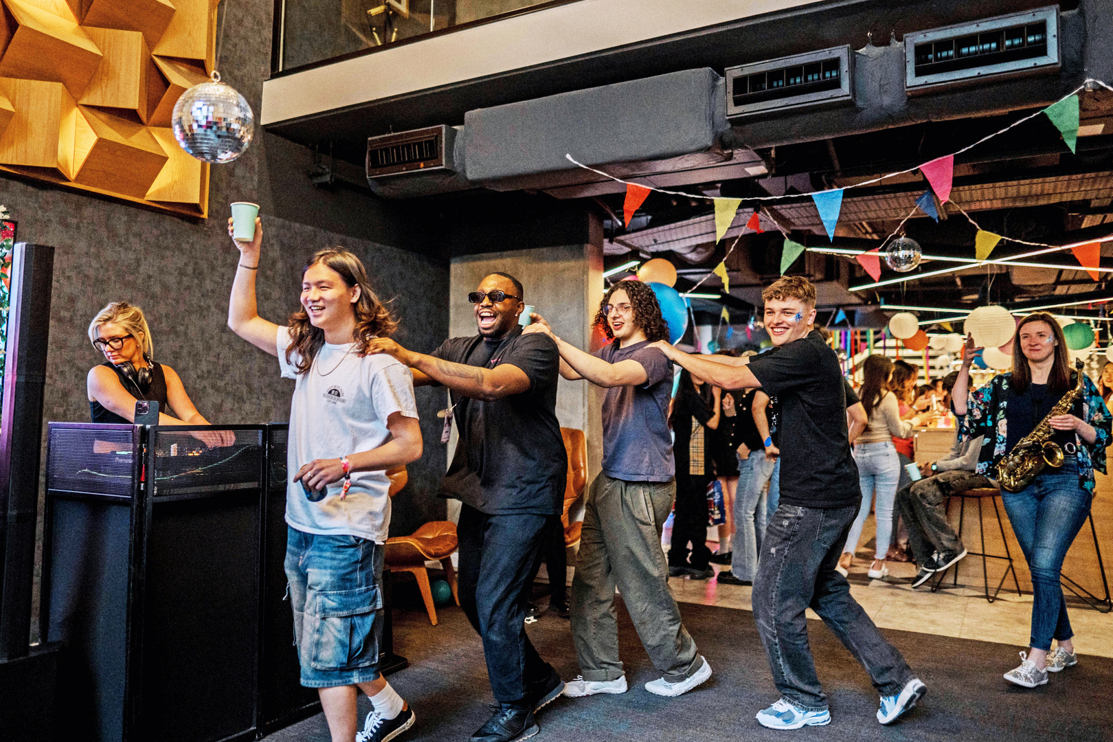 In a lively indoor setting, international students are dancing in a conga line under colorful streamers and a disco ball. One holds a cup while another plays the saxophone. The atmosphere is festive and joyful—the perfect place for connecting at Vita Student gatherings.