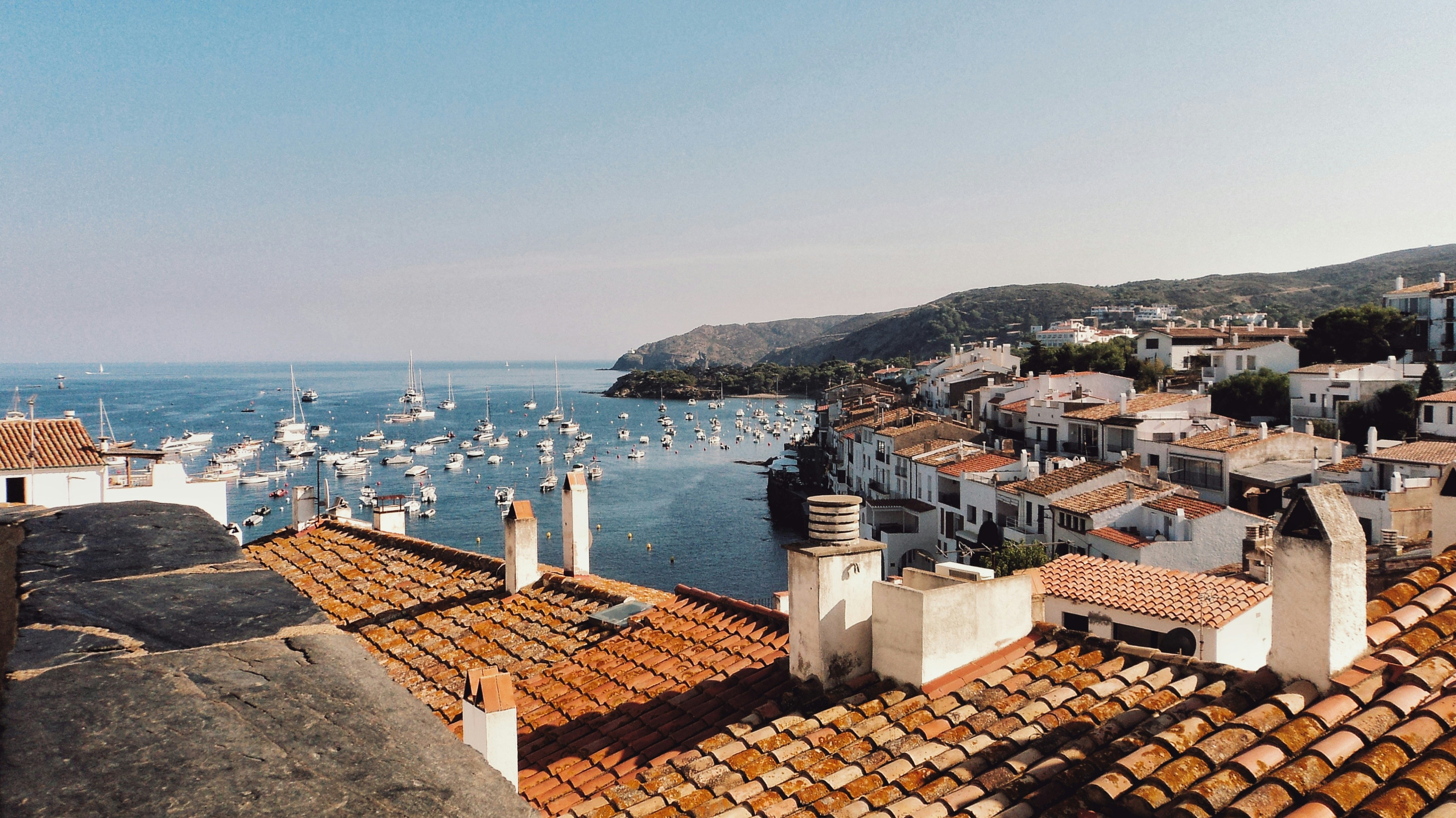 A coastal village with red-tiled roofs overlooks a serene bay filled with numerous small boats, ideal for day trips. Hills and greenery frame the background under a clear blue sky, reminiscent of picturesque scenes near Barcelona.