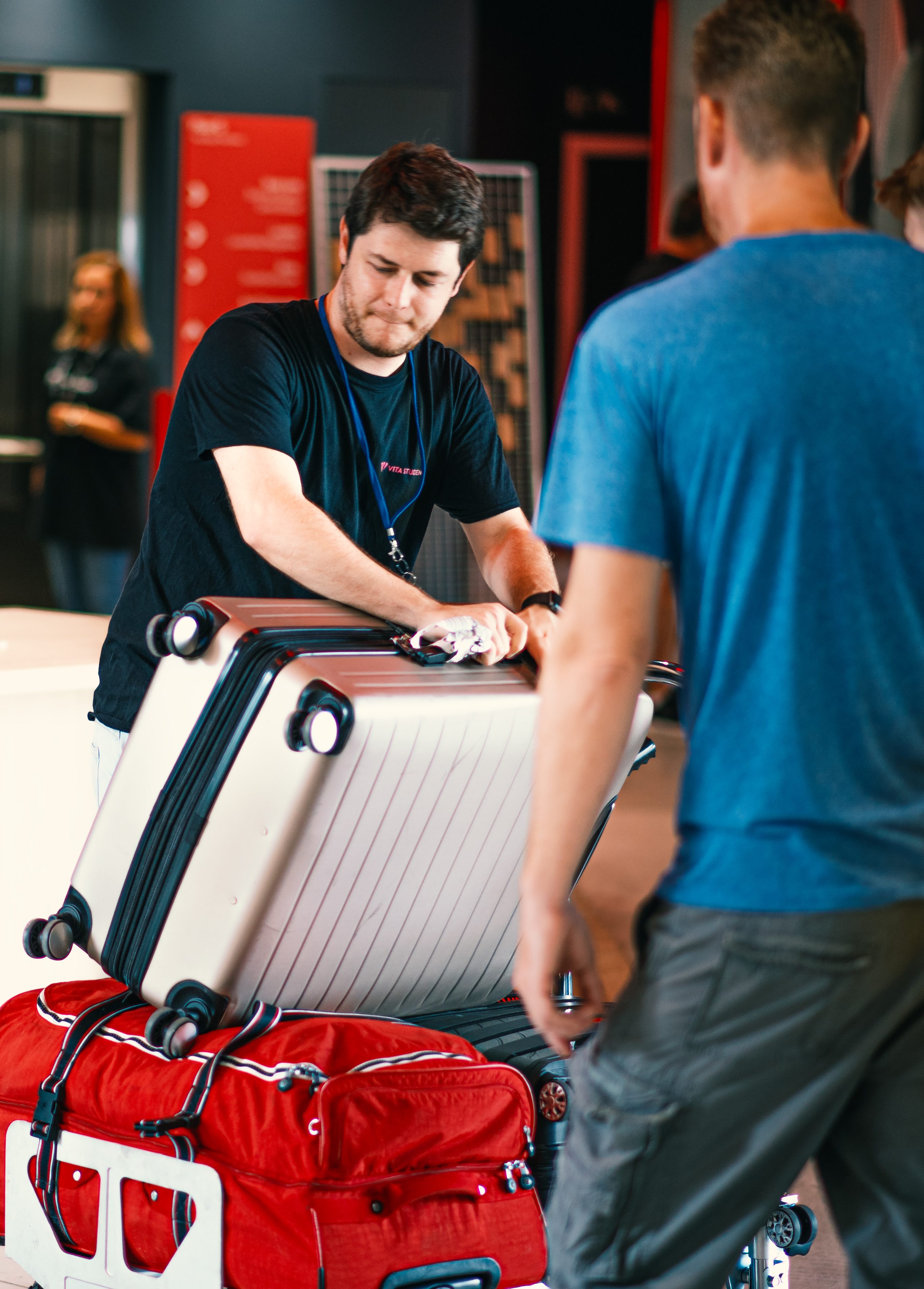 A man in a black shirt is handling a large white suitcase on a wheeled cart. A red duffel bag is also on the cart. Another person in a blue shirt is in the foreground, facing away. Indoor setting with blurred background.