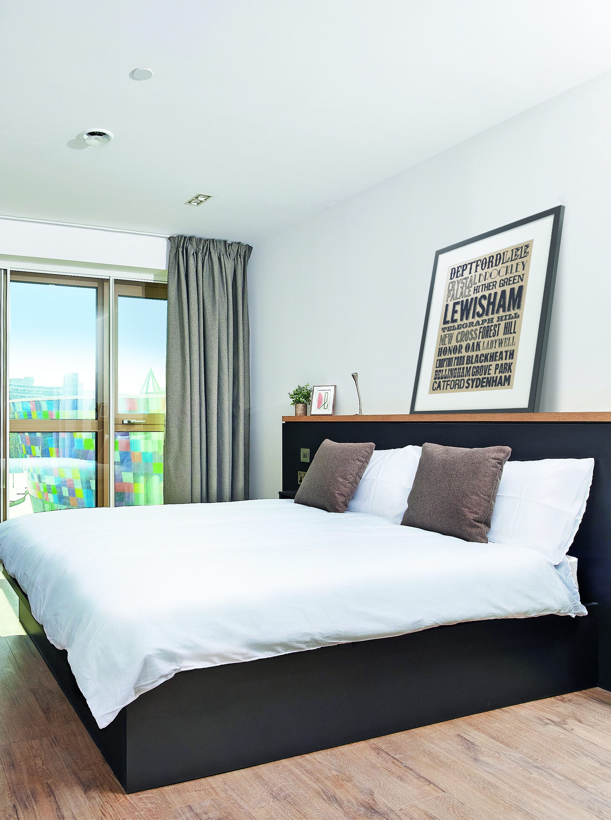 A modern bedroom with a large double bed featuring a white duvet and brown pillows. A framed poster is above the black headboard. A window with gray curtains opens to a balcony with a colorful view. Wooden flooring completes the room.