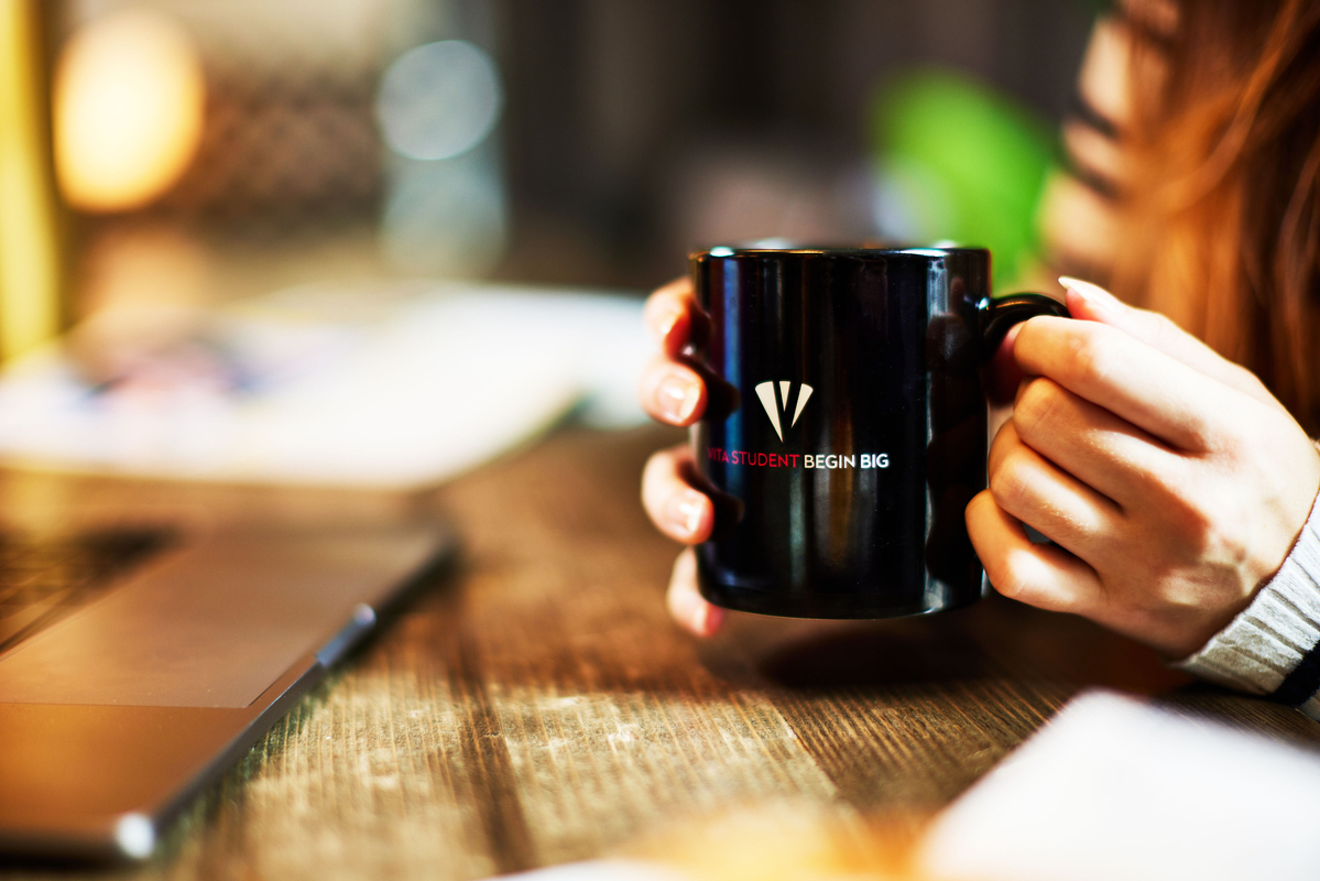 A person holding a black mug with the text "BEGIN BIG" on it, sitting at a wooden table next to a laptop. The background is slightly blurred, and the lighting is warm, creating a cozy atmosphere.