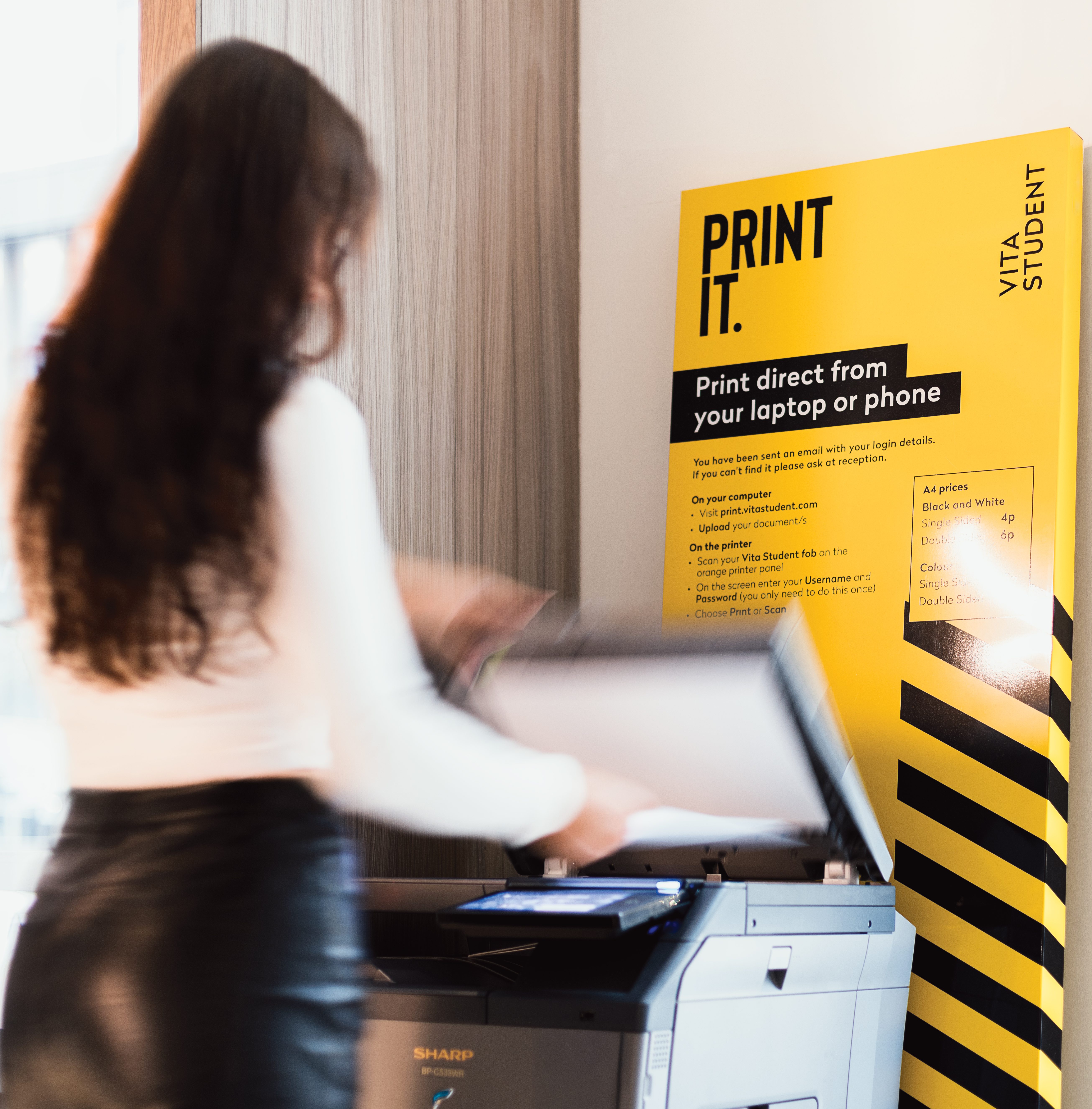 A person in a white shirt and black skirt uses an upgraded printer next to a bright yellow sign that reads, "PRINT IT. Print direct from your laptop or phone." The scene is indoors, featuring modern and minimalistic design, highlighting the benefits of seamless technology integration.