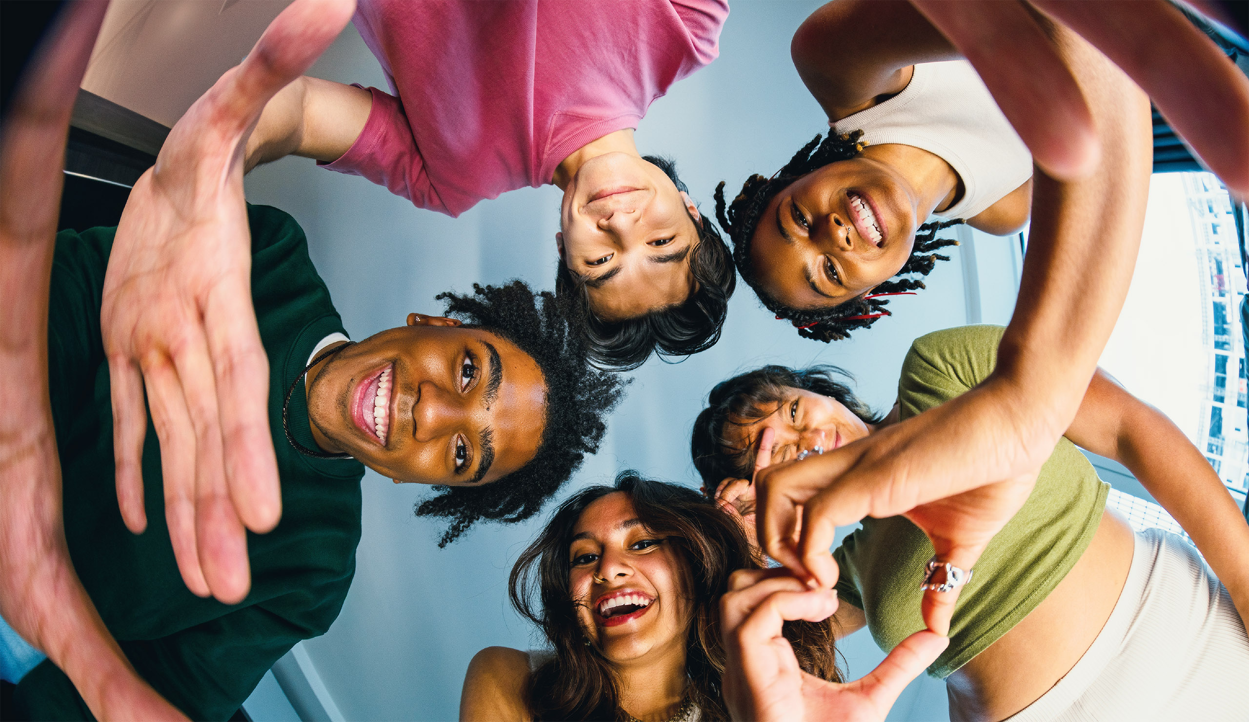 Five friends stand in a circle, facing downward towards the camera with joyful expressions. Indoors, their hands reach toward the lens, creating a dynamic and friendly atmosphere, as if to refer to an unspoken bond captured in this vibrant moment.