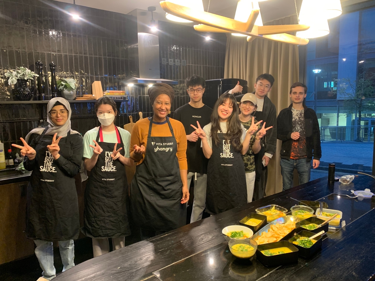 A diverse group of eight people stands in a modern kitchen, wearing aprons and smiling. Some are making peace signs. On the counter, there are dishes with prepared food. Overhead lights illuminate the scene.