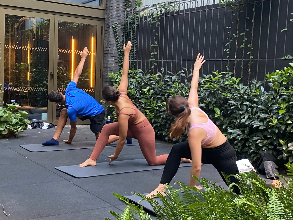 Three people practice yoga on mats outdoors, each in a low lunge with one arm raised. They are surrounded by greenery and positioned on a dark surface. A glass door and vine-covered wall are in the background.
