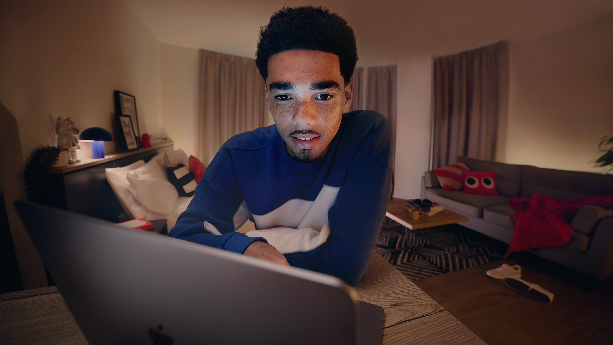 A person with short curly hair and freckles looks at a laptop screen intently, sitting on a bed in a dimly lit room. The room has a couch with a red blanket, curtains, and various decorative items around.