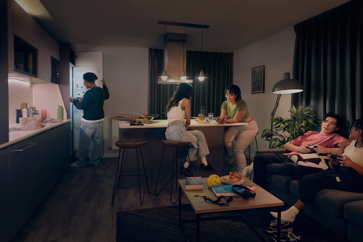 A cozy living room and kitchen scene with four people. One person is opening the fridge, two are sitting at a kitchen island chatting, and two are on a couch playing video games. The room is warmly lit with casual decor and snacks.