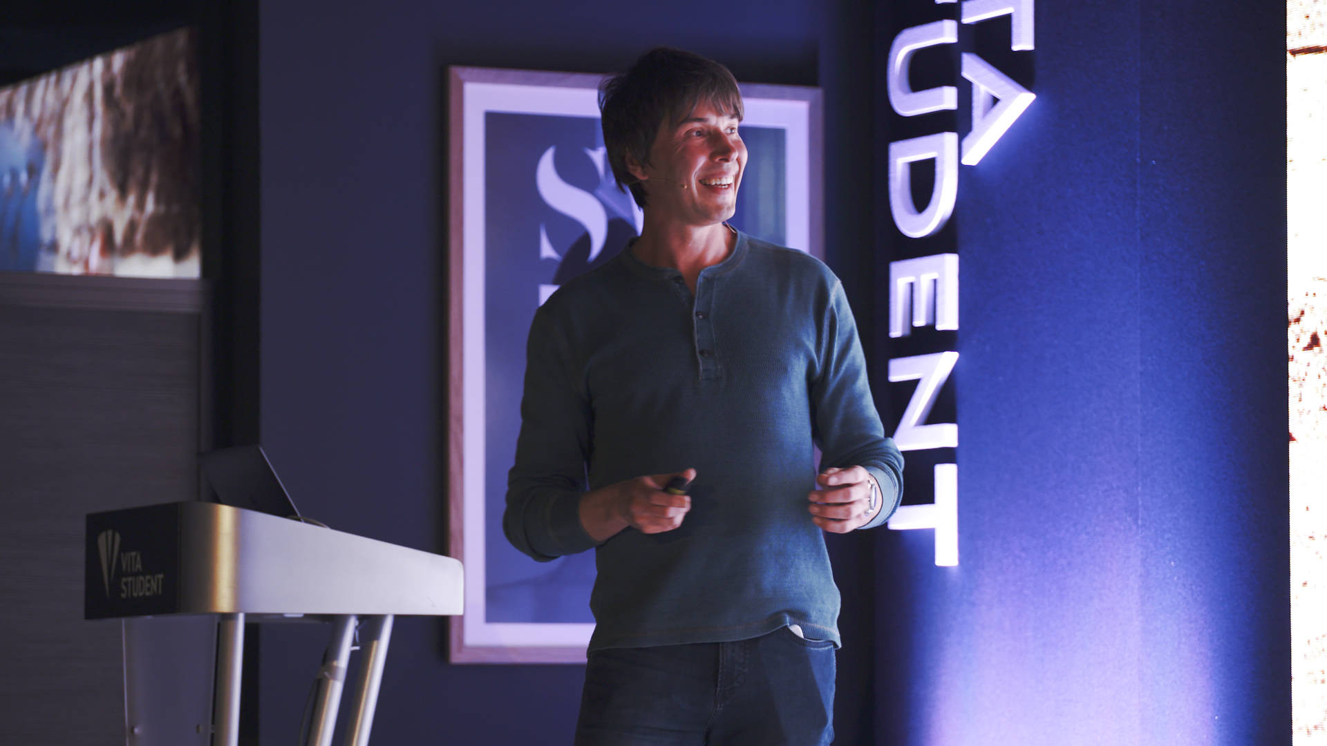 A person in a blue long-sleeve shirt stands smiling while giving a presentation. They are holding a clicker. The background features a framed picture and illuminated signage.