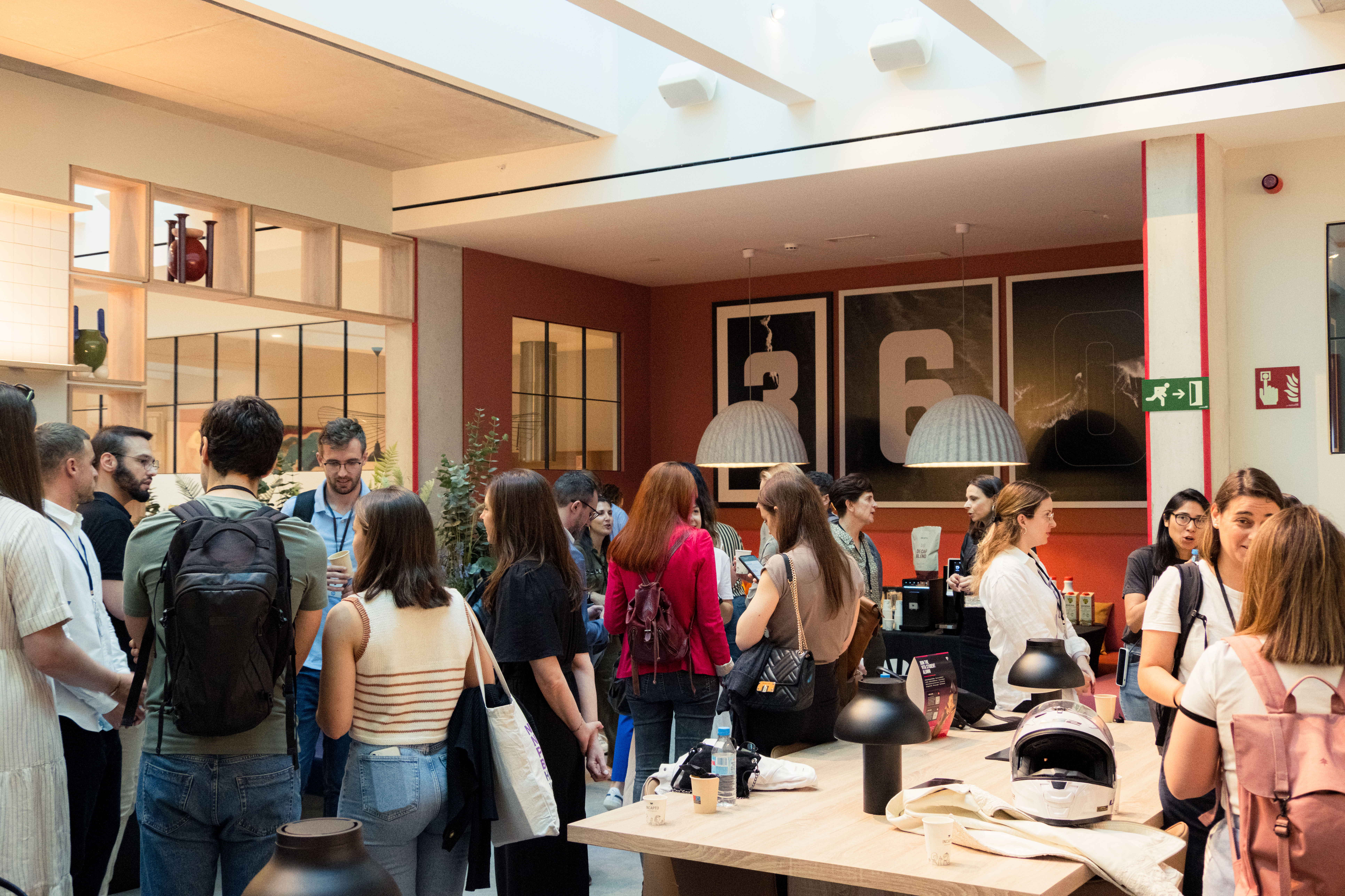 A group of people gather in a modern, well-lit room with a high ceiling. They are casually dressed and engaged in conversation. The room features contemporary decor, including pendant lights and framed artwork with numbers.