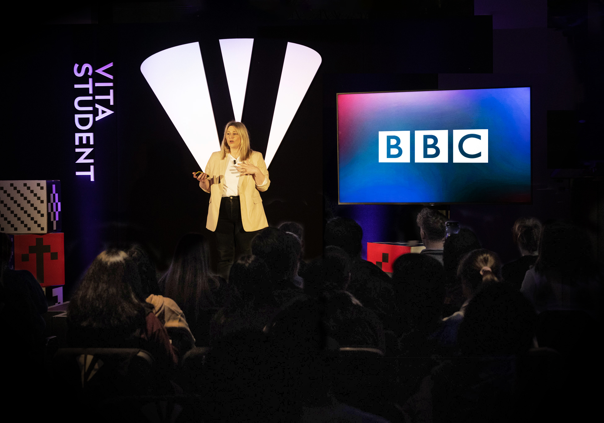 A person stands on stage speaking to an audience. They are holding a microphone and wearing a white blazer. Behind them is a large screen displaying the BBC logo. "Vita Student" is written on a vertical sign on the left.