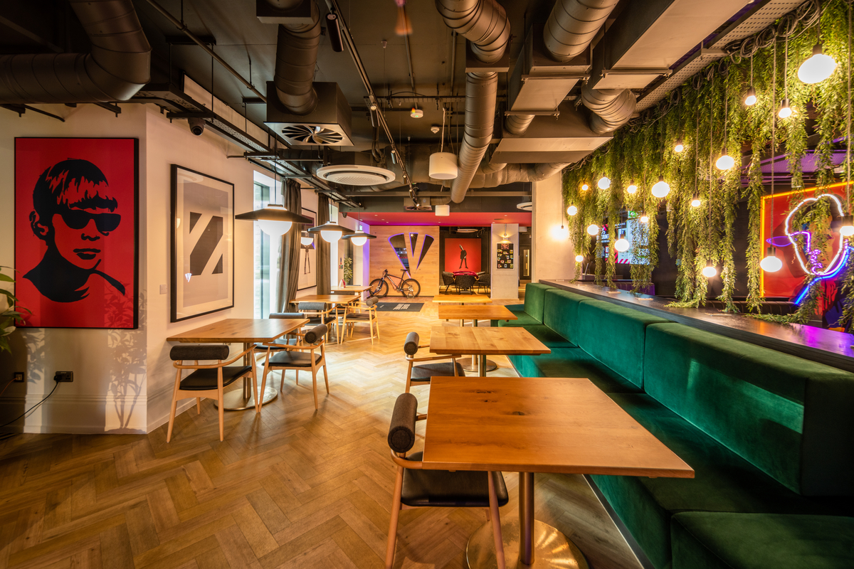 A modern cafe interior with wooden tables and green velvet seating. The ceiling features exposed pipes and hanging lights. The walls display vibrant art, including a portrait of a person in sunglasses. Indoor plants add greenery.
