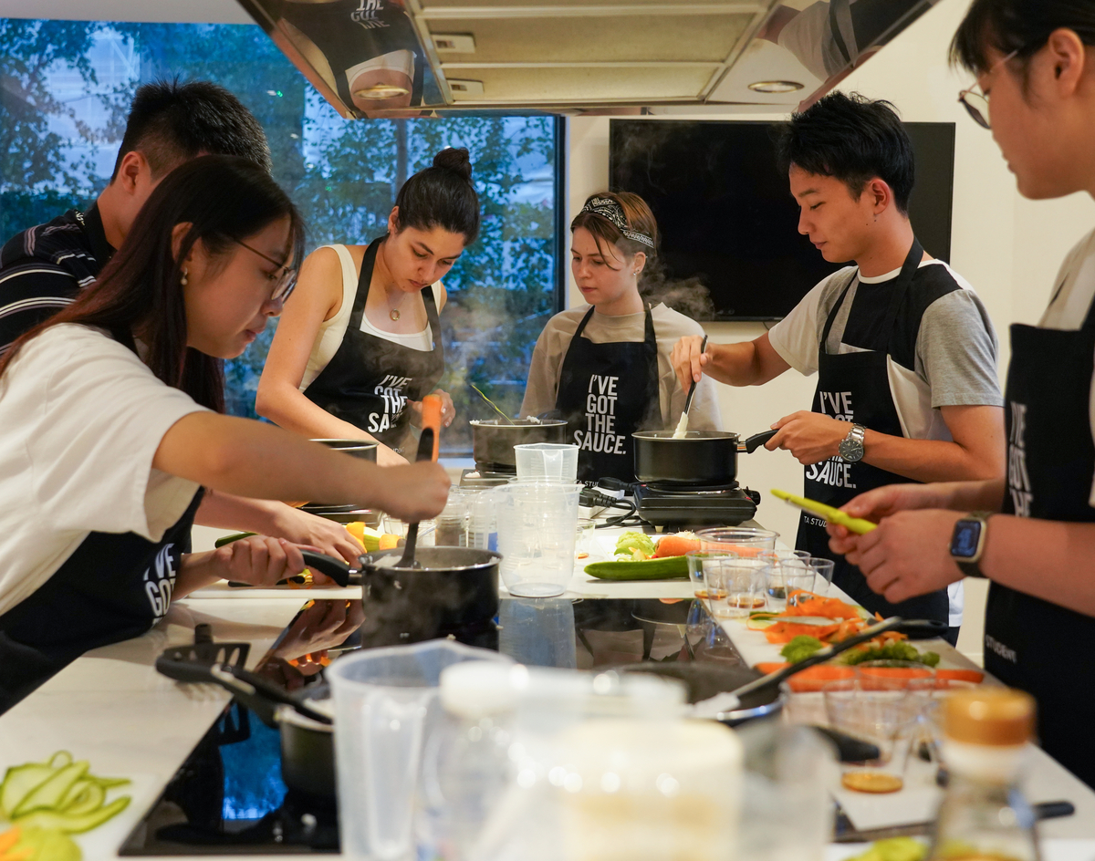 A group of young adults wearing aprons, cooking together in a modern kitchen. Various vegetables and kitchen utensils are visible on the counter. They appear focused and engaged in their culinary tasks.