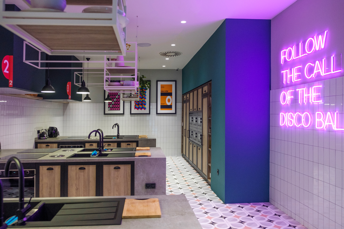Modern kitchen interior with wooden cabinetry and countertops, featuring sleek sinks and appliances. A neon sign on the wall reads "FOLLOW THE CALL OF THE DISCO BALL." The room is decorated with colorful art and patterned floor tiles.