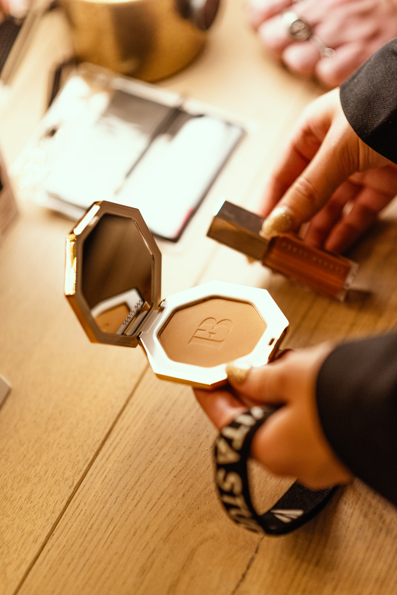 A person holds a compact powder case with a mirror and logo, alongside a rectangular makeup item. The items are on a wooden surface.