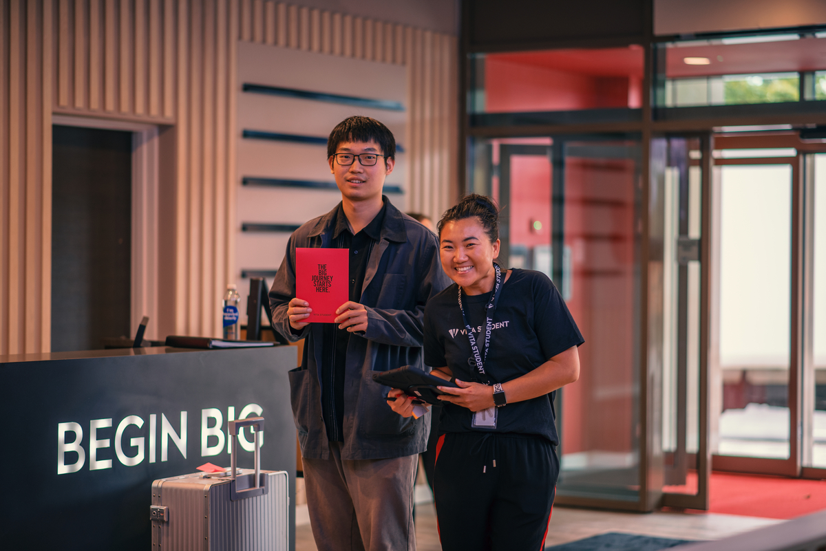 Two people stand smiling in a modern lobby. The person on the left holds a red booklet, and the person on the right holds a tablet. A large sign on a desk reads "BEGIN BIG." Bright lighting creates a welcoming atmosphere.