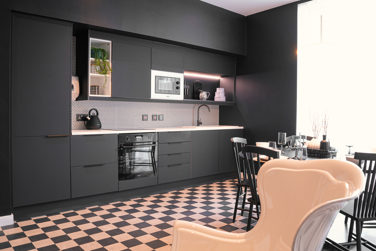 A modern kitchen with matte black cabinets, a checkered black and white floor, and a tiled backsplash. Appliances include a built-in microwave and oven. A white countertop holds a black kettle. A dining table with black chairs is in the foreground.