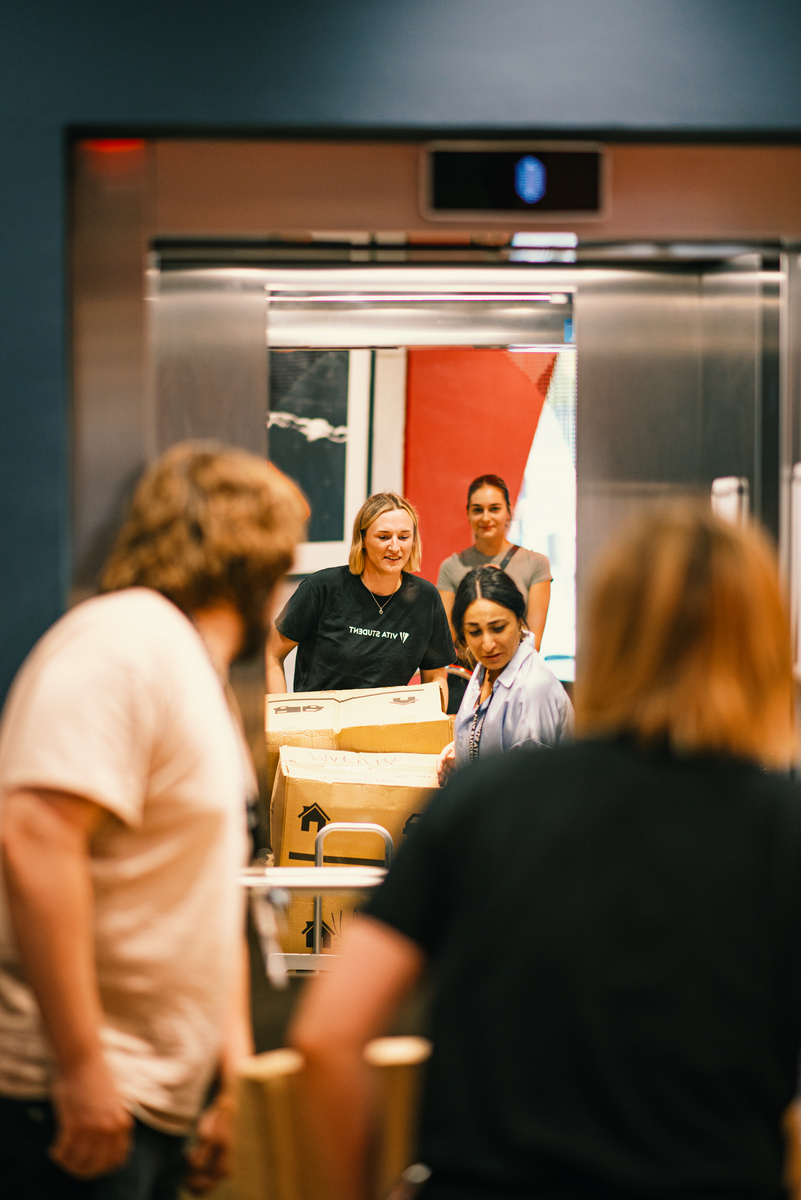 A group of people are unloading large cardboard boxes from an elevator. They are working together, with one person inside the elevator and others outside, ready to assist. The scene is set in a building with blue walls and vibrant artwork.
