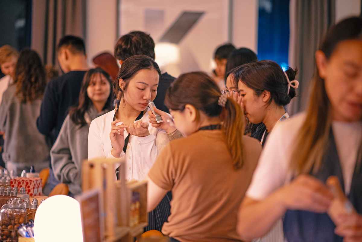 A group of people in a bustling indoor market, sampling and smelling products. A woman in the foreground smells a sample on her wrist while others engage with items on display. Warm lighting creates a cozy atmosphere.