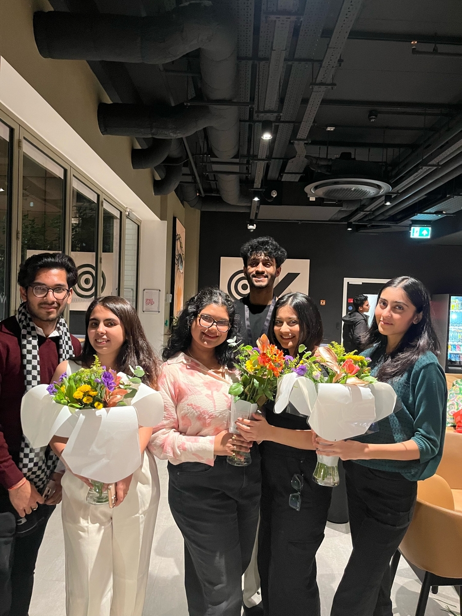 A group of six people indoors, smiling and holding bouquets of colorful flowers. They are standing in a line, with bright indoor lighting and a modern interior design with black ceilings and large windows.