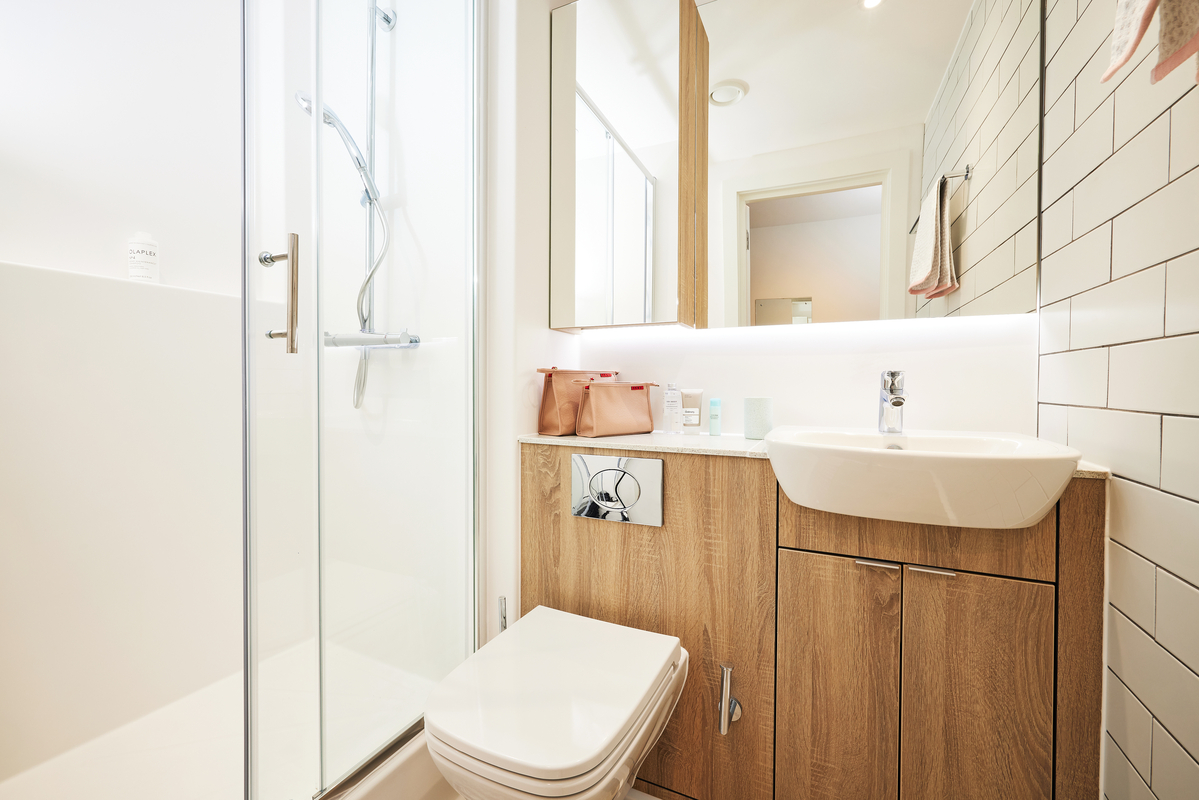 Modern bathroom with beige tiles, a glass shower enclosure on the left, a wooden vanity unit with a white sink, and a toilet. There are toiletries on the counter and a mirrored cabinet above. Walls have white subway tiles.