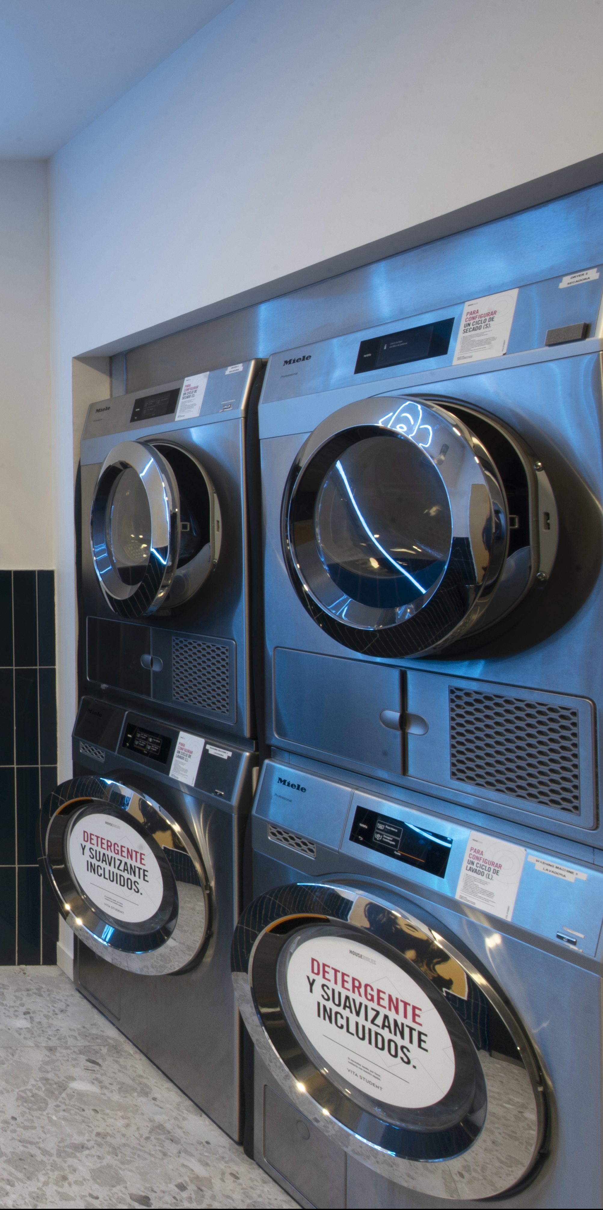 A laundromat with four stainless steel washing machines stacked in pairs boasts upgraded benefits: each machine displays a sign reading "Detergente y Suavizante Incluidos," indicating detergent and softener are included. The floor is tiled, and the walls are light-colored.