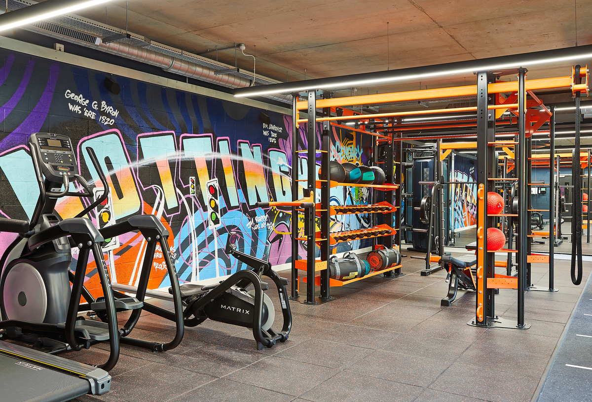 A modern gym with colorful graffiti on the walls. It features exercise equipment like treadmills, weight racks, and a functional training rig. The space has bright lighting and a mix of black and orange accents.