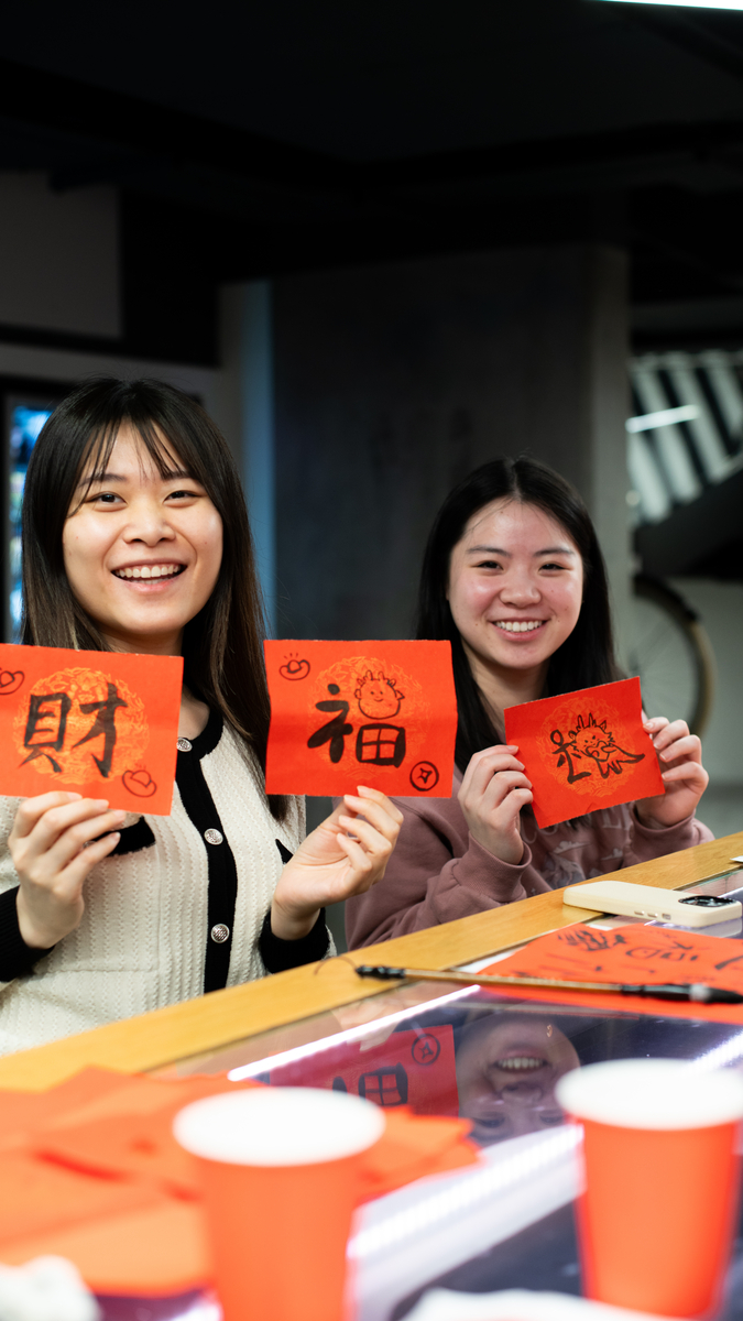 Two people smiling and holding red paper squares with handwritten calligraphy. They are sitting at a table with more red papers and cups around. The setting appears to be festive, possibly related to a cultural celebration.