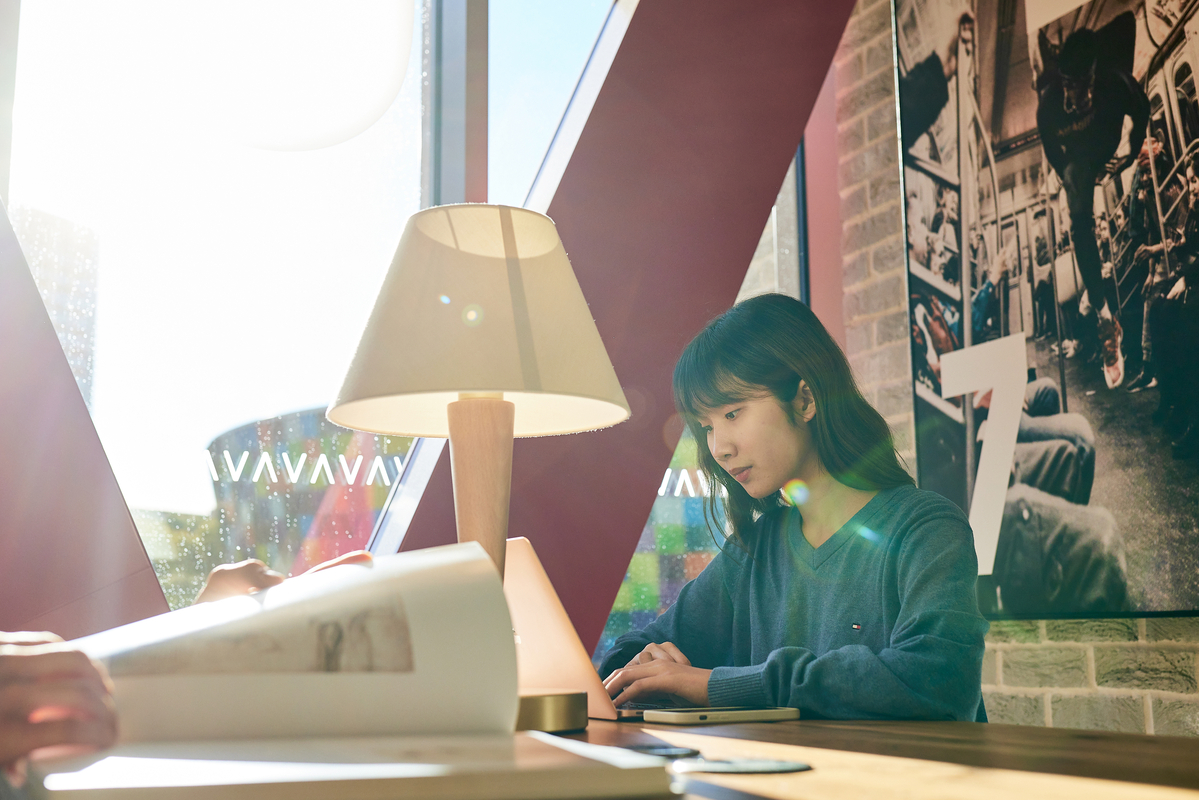 A woman in a blue sweater sits at a table, writing on a notepad, with bright sunlight streaming through a large window. A lamp and an open book are on the table, and a wall with photos is in the background.