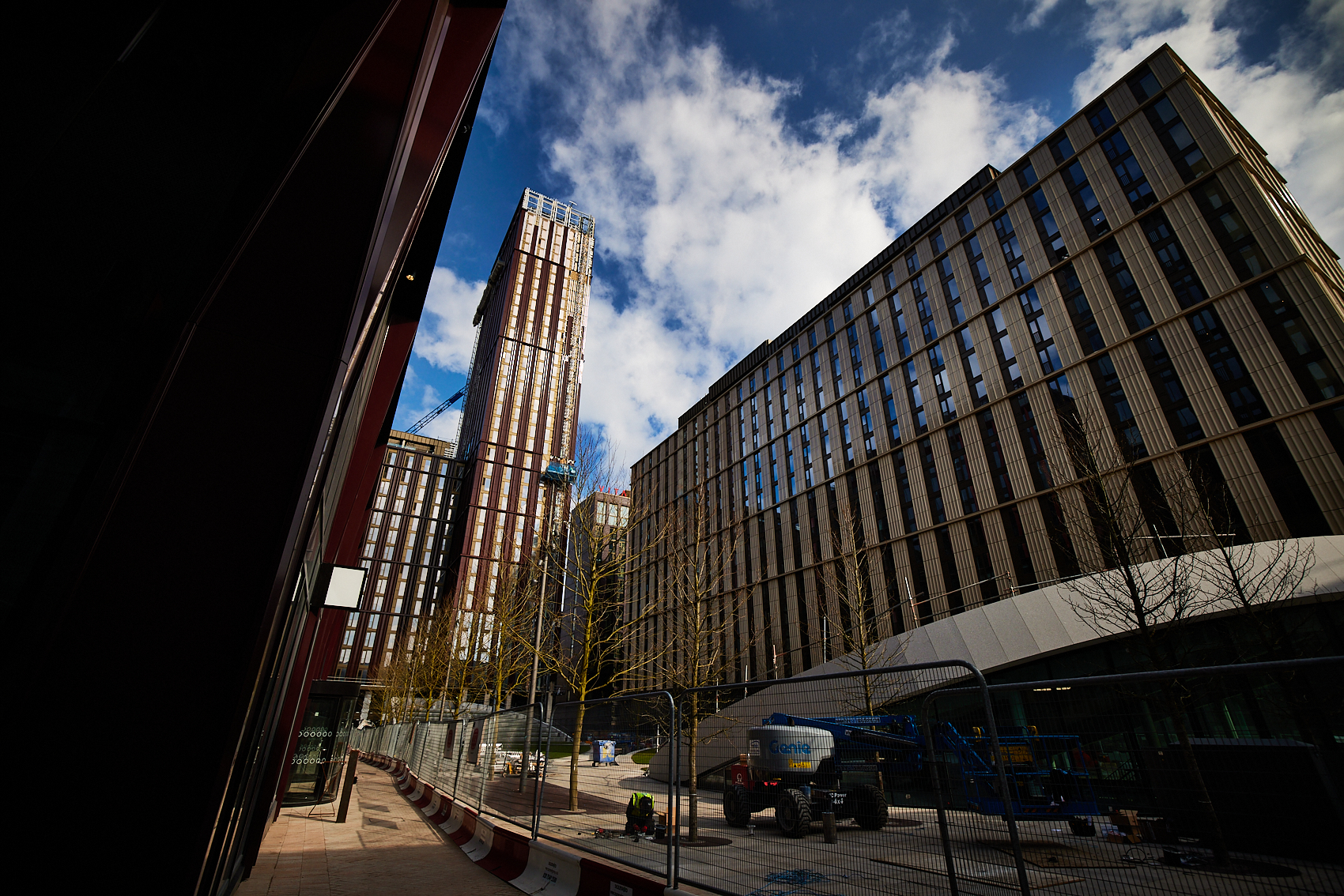 Tall, modern buildings with glass and metal facades rise under a partly cloudy sky. Student halls are nestled among the structures. A construction area is visible at street level with a truck and barriers, while leafless trees line the bustling street.
