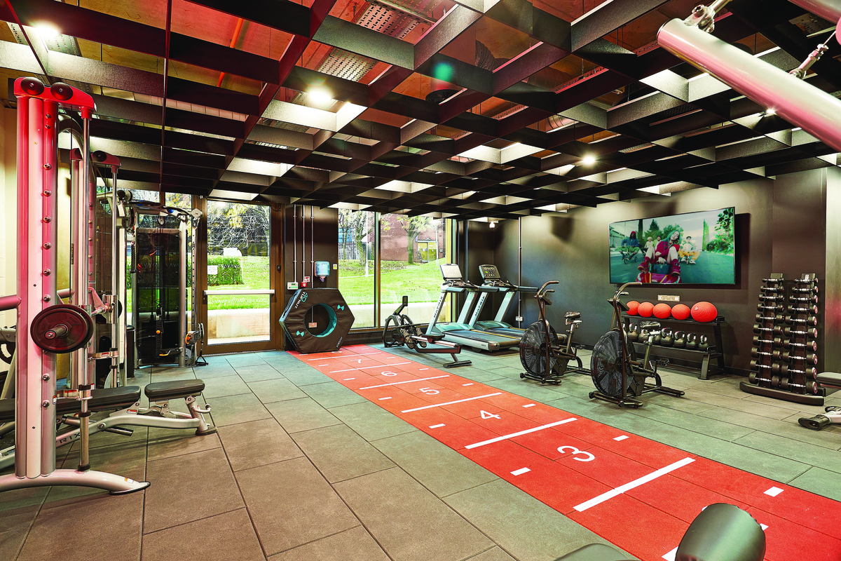 Modern gym with various equipment including treadmills, stationary bikes, weight machines, free weights, and a rack of exercise balls. The floor has a red and white marked track, and large windows provide natural light.