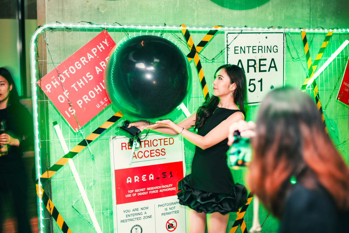 A woman in a black dress stands by a wall with Area 51-themed signs, including "Photography of This Area is Prohibited" and "Restricted Access." She holds a large black balloon. The scene is lit with green lighting.