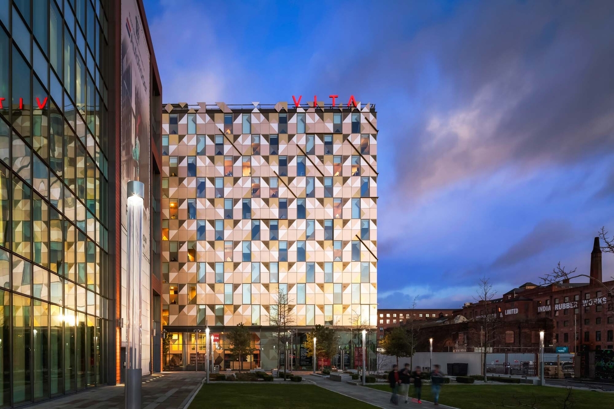 Modern building with a geometric facade featuring triangular patterns at dusk. The structure has a glass-fronted entrance and the word "VITA" visible in red letters on top. People walk on a pathway in the landscaped area in the foreground.