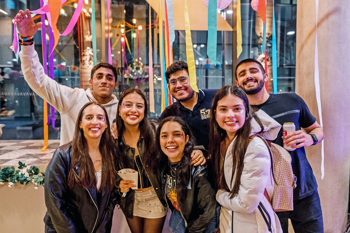 A group of seven people smiling at a party. They are standing close together with colorful streamers in the background. Two individuals are holding drinks. Everyone is wearing casual, festive clothing.