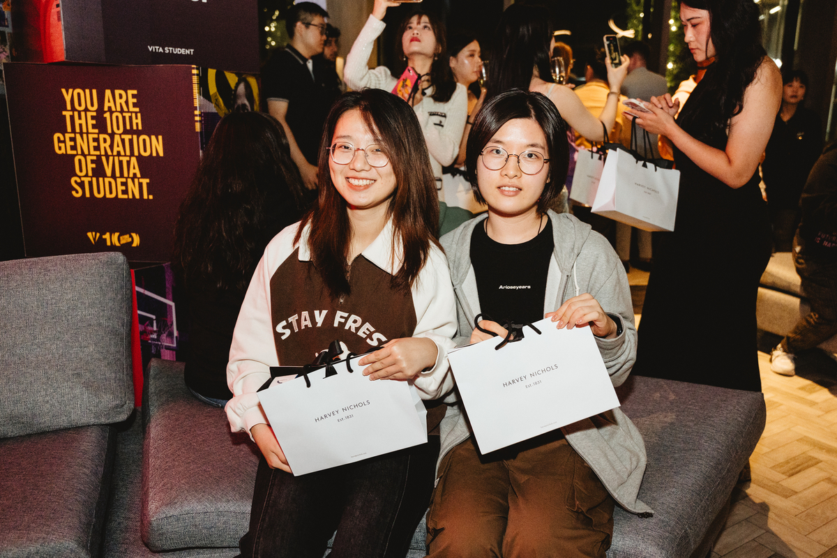 Two people sitting on a couch, smiling and holding Harvey Nichols shopping bags. A group of people stand in the background, some holding phones. A sign reads, "YOU ARE THE 10TH GENERATION OF VITA STUDENT." The scene is well-lit and lively.