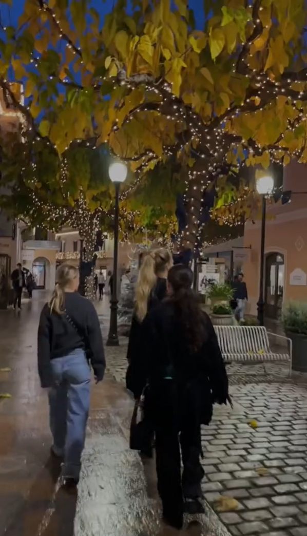 Three people walk through a charming street decorated with fairy lights wrapped around trees. The cobblestone path glistens, reflecting the soft evening glow. The scene evokes a cozy, festive atmosphere.