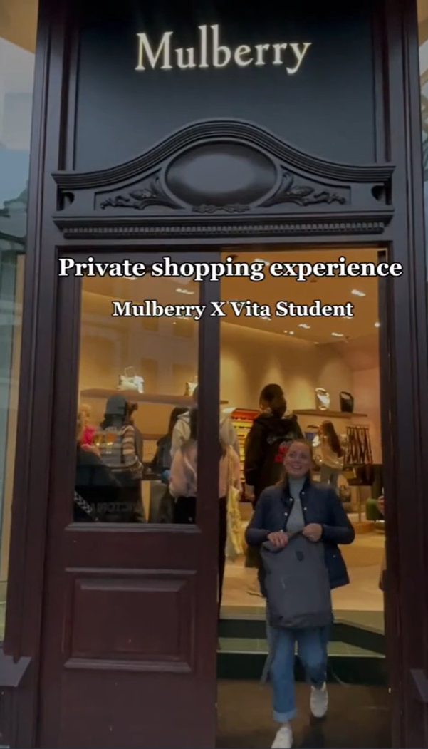 A person exits a Mulberry store holding a bag, with a sign above reading "Private shopping experience Mulberry X Vita Student." The interior shows other people browsing. The storefront is elegant and wooden.