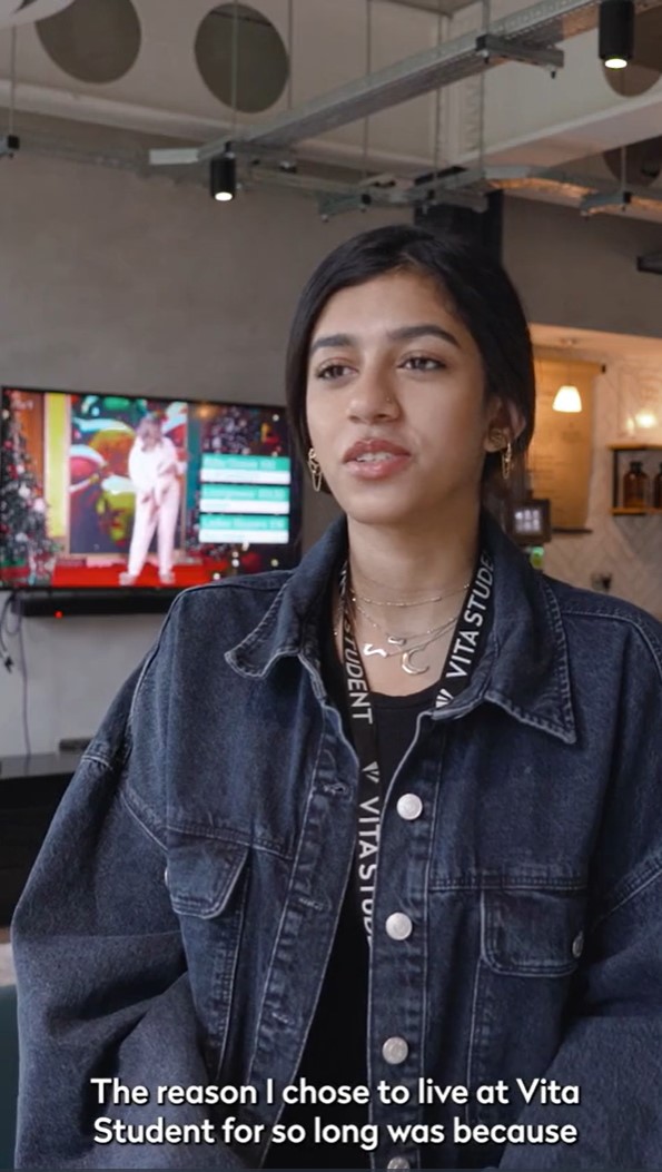 A person wearing a denim jacket stands indoors, speaking. In the background, a television shows a colorful image. The text at the bottom reads: "The reason I chose to live at Vita Student for so long was because of their vibrant international student community.