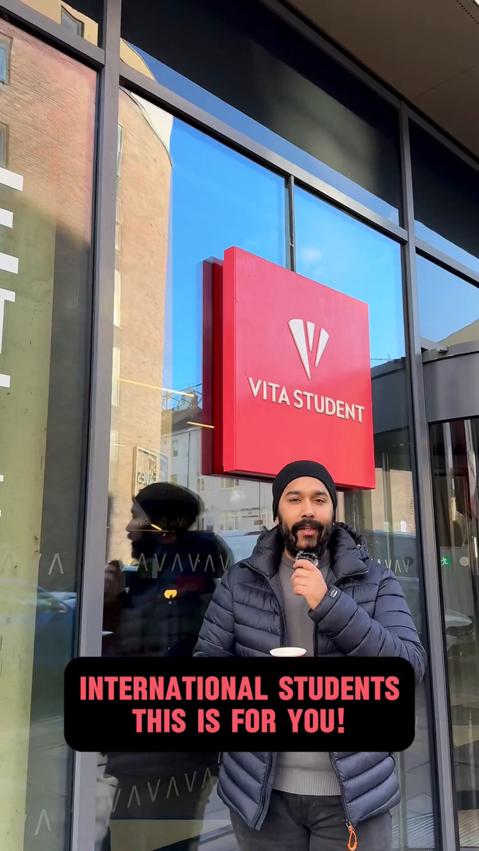 A person stands in front of the Vita Student building, microphone in hand. The glass facade reflects nearby structures, creating a vibrant scene. A text overlay reads, "Calling all international students—this is for you!.