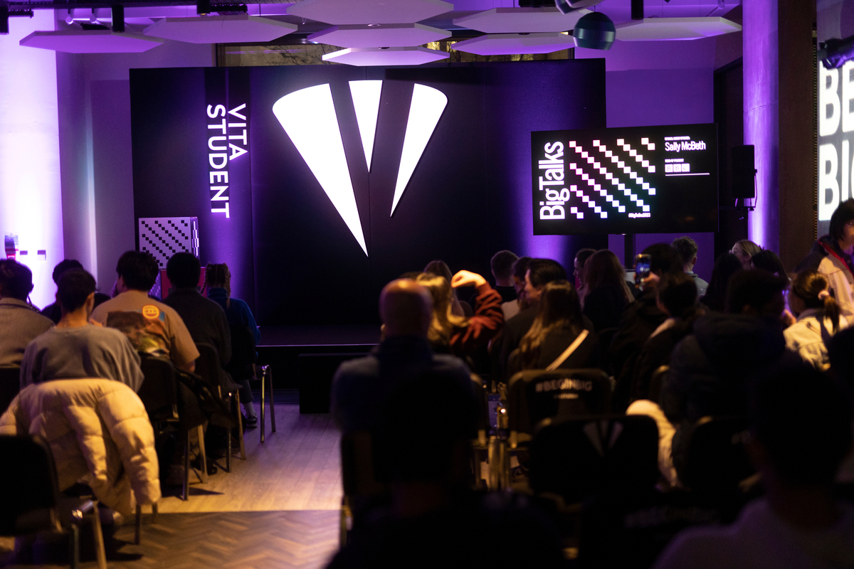 Audience seated in a dimly lit venue with purple lighting, facing a stage displaying "VITA STUDENT" and "Big Talks." A presentation is about to begin.