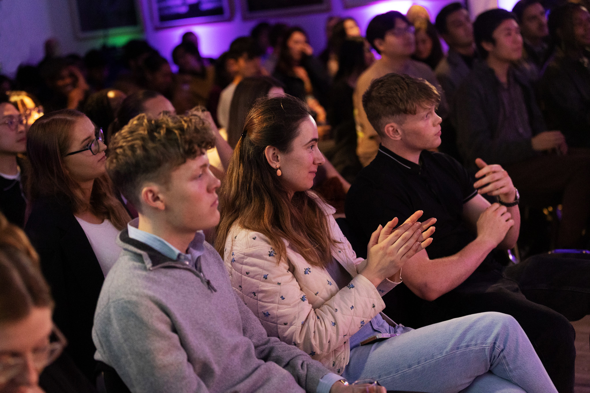 A group of people sitting closely together in a dimly lit room. They appear to be focused on something off-camera, with some individuals clapping. The background is softly illuminated with purple lighting.