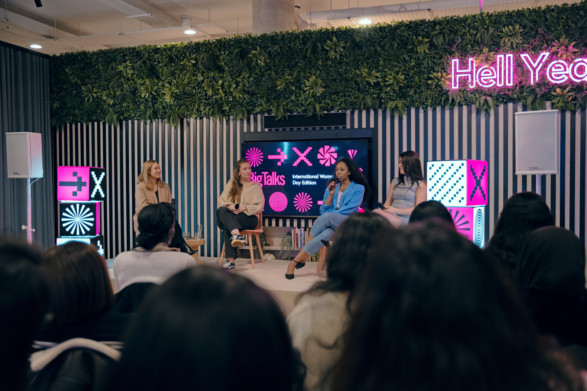 A panel of four women sits on a stage with a neon sign that reads "Hell Yea" and digital screens displaying "X Talks International Women's Day Edition." An audience watches in the foreground. The backdrop includes vertical stripes and greenery.